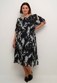 Zwart-wit geplooide jurk met een losse pasvorm, korte gerimpelde mouwen en een strikdetail in de taille. Schoenen zijn zwarte puntige ballerina's.