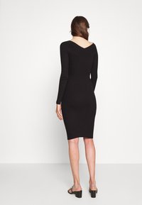 Černé žebrované bodycon šaty s dlouhými rukávy, designem V-zpět, délka do poloviny lýtek a přiléhavý střih. Nosené s černými sandály na blokovém podpatku.