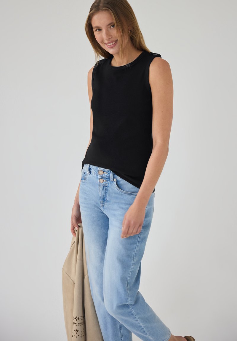 Jeune femme souriante, portant un haut noir sans manches et un jean bleu clair, tenant une veste beige dans la main gauche, debout devant un fond gris uni.