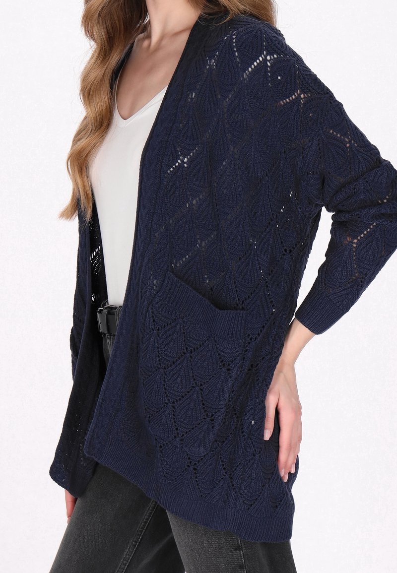 Cardigan en maille bleu marine à ouverture devant, avec un motif feuille texturé, manches longues et une poche avant. Porté sur un haut blanc.