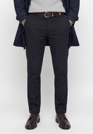Homme portant un pantalon bleu marine foncé avec une ceinture en cuir marron, des chaussures habillées marron, un pull gris et un manteau bleu marine, les mains dans les poches.