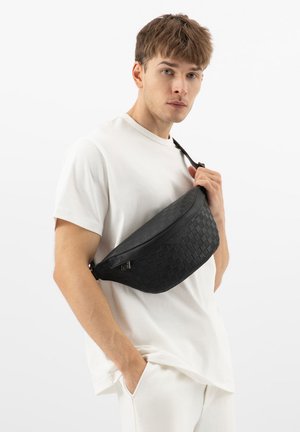 Młody mężczyzna w białym stroju, trzymający czarną, fakturowaną torbę na ramię jedną ręką, patrzący w stronę aparatu na jednolitym białym tle.