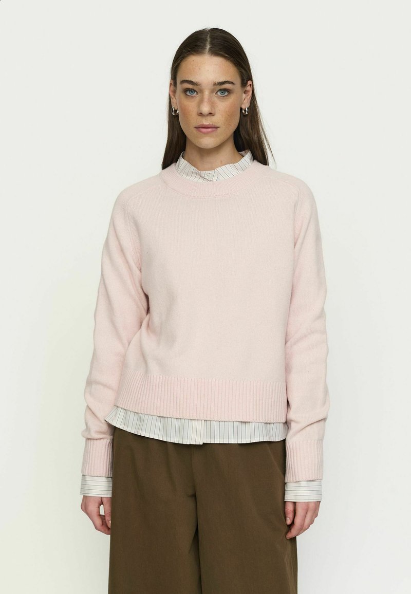 Femme aux cheveux bruns raides portant un pull rose pâle sur une chemise rayée et un pantalon marron, debout devant un fond uni clair.