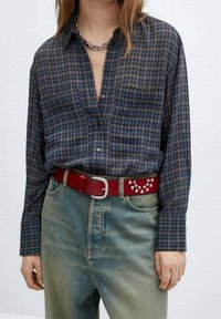 Chemise à carreaux boutonnée en bleu marine, marron et beige avec des poches poitrine ; associée à un jean bleu délavé et une ceinture rouge ornée de clous.