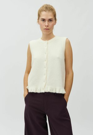Femme aux cheveux blonds portant un gilet en laine blanc sans manches boutonné et un pantalon large foncé, debout les mains dans les poches devant un fond uni.