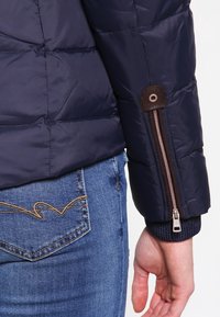 Veste matelassée bleu marine avec un design matelassé. Présente un accent marron au niveau du poignet et une ouverture zippée sur la manche. Portée avec un jean bleu.