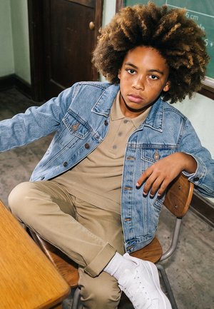 Junge mit lockigem Haar, der eine Jeansjacke und khakifarbene Kleidung trägt, sitzt auf einem Stuhl im Klassenzimmer mit übereinandergeschlagenen Beinen und einem Arm auf der Rückenlehne abgestützt.