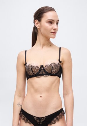 Frau mit langen braunen Haaren in einem niedrigen Pferdeschwanz, die einen schwarzen Spitzen-BH und passende Spitzenunterwäsche trägt und nach rechts blickt, vor einem schlichten Hintergrund.