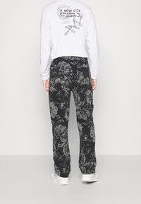 Long-sleeve white shirt features graphic print on back. Black pants with a floral pattern, soft texture, and a relaxed fit. White sneakers.
