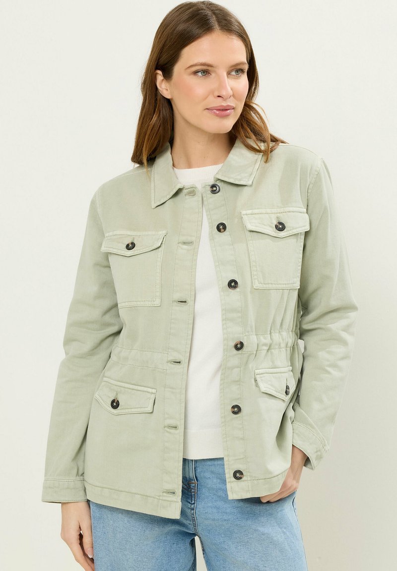 Frau trägt eine hellgrüne Jacke mit Knöpfen und vier Klappentaschen über einem weißen Oberteil und blauen Jeans, schaut nach rechts mit einem neutralen Gesichtsausdruck.