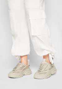 Beżowe sneakersy z siateczkowymi i solidnymi wstawkami, z grubą podeszwą i sznurowanym zapięciem. Standardowy gumowy bieżnik i pętla na pięcie ułatwiające zakładanie.