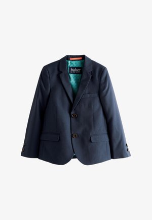 Junger Junge mit blondem Haar, der einen marineblauen Anzug, weißes Hemd und weiße Turnschuhe trägt, steht vor einem schlichten hellen Hintergrund.