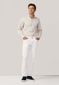 Camicia button-up di un beige chiaro con tasca sul petto, abbinata a jeans bianchi e sneakers nere. Design semplice, texture liscia e forma aderente.