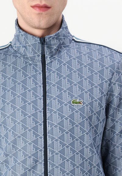 Veste zippée bleue avec un motif géométrique, col haut et bandes latérales contrastées. Présente un petit logo de crocodile vert sur la poitrine.