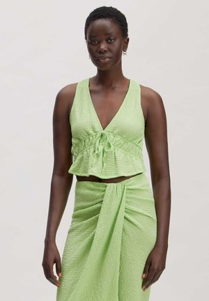Woman wearing textured light green sleeveless crop top with tie front and matching ruched skirt, standing against plain light background.