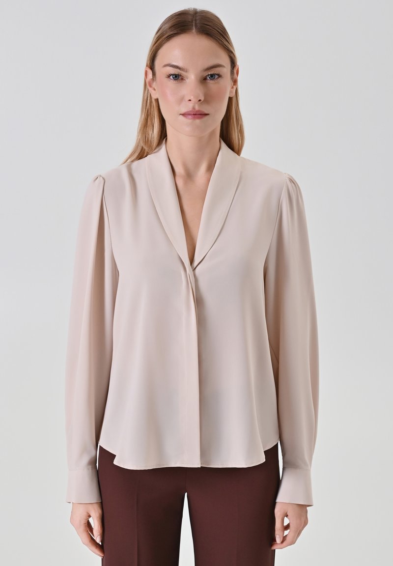 Helle beige Bluse mit Schalkragen, langen Ärmeln und glatter Textur. Verfügt über einen dezenten Fall und eine frontale Verschluss.