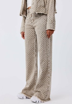 Jeans de pierna ancha de color beige con un acabado texturizado, que presentan bordes deshilachados y un ajuste relajado, combinados con zapatillas blancas.