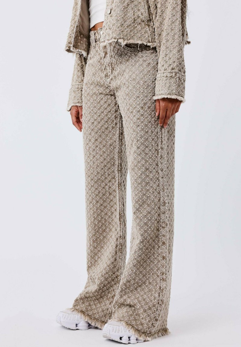 Jeans de pierna ancha de color beige con un acabado texturizado, que presentan bordes deshilachados y un ajuste relajado, combinados con zapatillas blancas.
