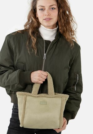 Green bomber jacket, off-white turtleneck, holding a beige faux shearling tote bag with a textured, fluffy edge and fabric label.