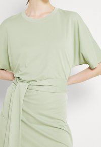Jasnozielona sukienka T-shirt z krótkim rękawem, z detalem w postaci związanej talii. Miękka, gładka tkanina z luźnym krojem i minimalistycznym designem.