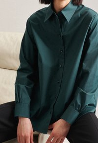 Mörkgrön skjorta med knappar, tillverkad av slät tyg, med klassisk krage, långa ärmar med åtsittande manschetter och svarta knappar.