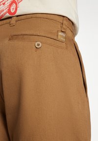 Pantalon en toile marron avec une finition texturée, doté d'une poche arrière unique avec fermeture à bouton et d'une étiquette de marque cousue au-dessus.