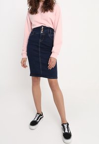 Jeanskjol i mörkblått med hög midja och femknappsstängning, kombinerad med en rosa sweatshirt och svarta sneakers.