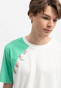 Korte mouw T-shirt met een groene linkerkant, een witte rechterkant en gedetailleerde illustraties van mensen die verschillende activiteiten uitvoeren nabij de halslijn.