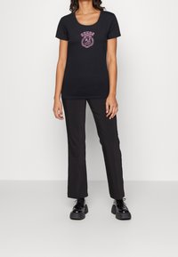 Camiseta negra de manga corta con escote en forma de bote, presenta el logo bordado en rosa "GUESS" y detalles florales. Combinada con pantalones de campana negros.