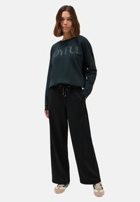 Dunkelgrüner Sweatshirt mit der Aufschrift "JOYFUL", kombiniert mit lockeren schwarzen Joggern und beigen Sneakers. Lässig, weicher Stoff, entspannte Passform.