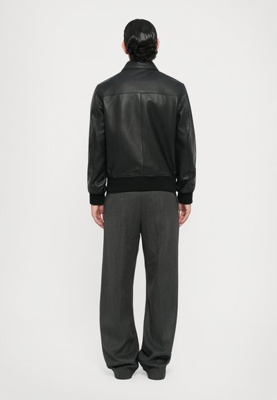 Veste en cuir noir avec des poignets et un ourlet côtelés, dotée d'un col classique et d'une texture lisse; associée à un pantalon gris ample.