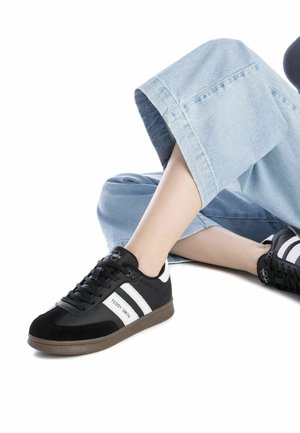 Zapatilla en blanco y negro con superficies de ante y cuero liso, que cuenta con una suela de goma y dos franjas blancas a los lados.