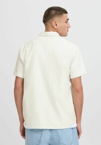 Chemise à manches courtes en tissu texturé blanc cassé, avec un col, un ourlet droit et un motif subtil, portée sur un jean bleu clair.