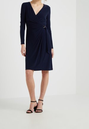 Femme portant une robe portefeuille bleu marine à manches longues avec un nœud sur le côté et des sandales noires à talons avec bride à la cheville, debout sur un sol blanc.