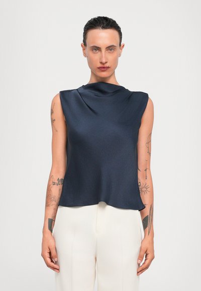 Top sans manches de couleur marine en tissu lisse, avec un col drapé et une coupe décontractée, associé à un pantalon de couleur claire.