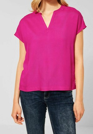 Kobieta w jasnoróżowej bluzce z krótkimi rękawami i dekoltem w serek oraz w ciemnoniebieskich dżinsach na jednolitym tle.