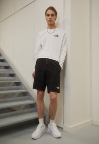 Chemise blanche à manches longues avec logo, shorts noirs et baskets blanches. Un homme se tient contre un mur, avec des escaliers visibles en arrière-plan.