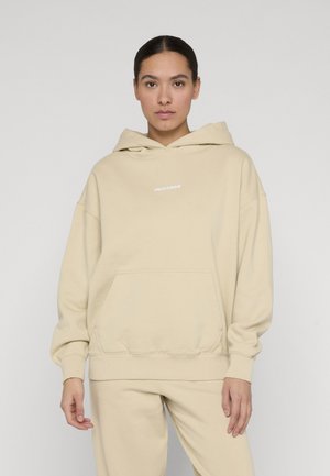 Frau trägt einen beigen Kapuzenpullover und passende Hose, steht vor einem schlichten hellgrauen Hintergrund.