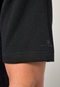 Manica di T-shirt nera in cotone con una texture morbida. Presenta un orlo cucito e un sottile logo "JP AWARE" in rilievo vicino al polsino.