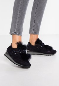 Navy suede sneakers featuring faux fur accents, floral appliqués, and a black rubber sole. Grey cropped jeans are paired with them.