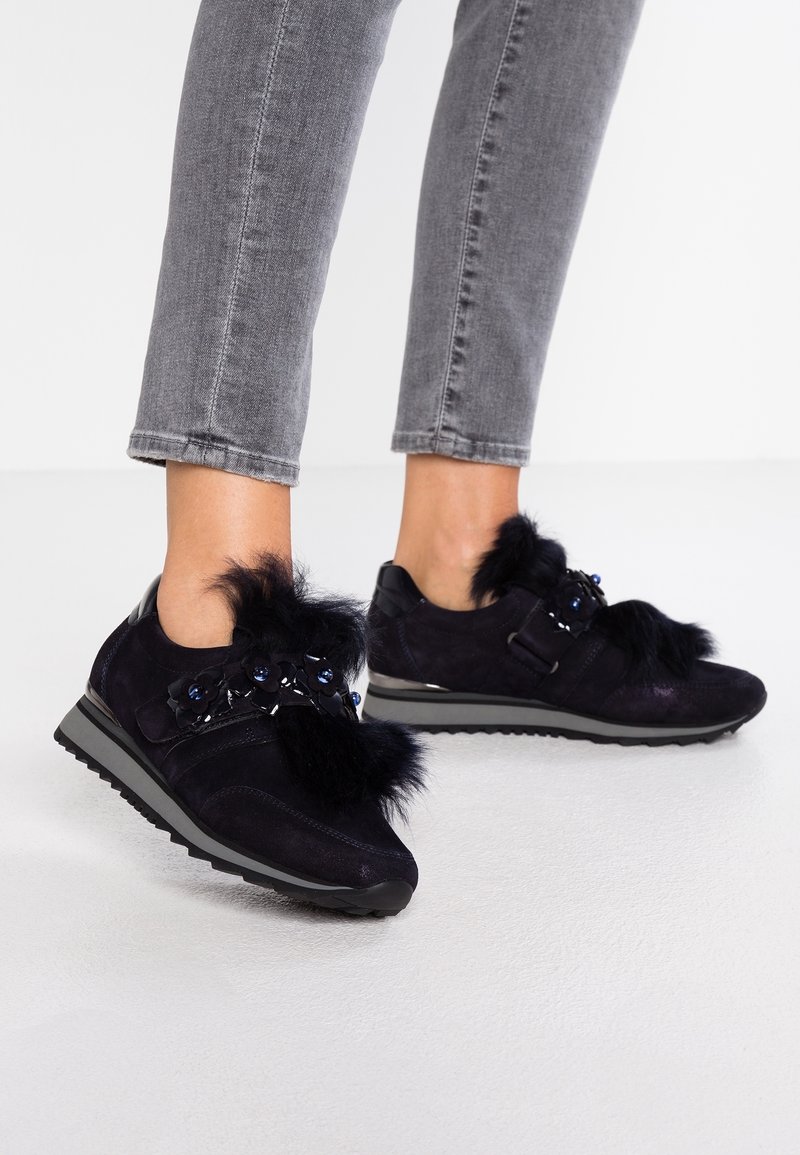 Navy suede sneakers featuring faux fur accents, floral appliqués, and a black rubber sole. Grey cropped jeans are paired with them.