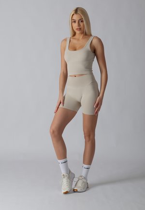 Mujer joven de pie, con top corto sin mangas beige y pantalones cortos ajustados a juego con calcetines blancos y zapatillas deportivas sobre fondo gris liso.
