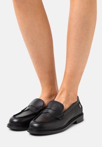 Mustat nahkaiset loafersit matalalla korolla, joissa on ommeltu muotoilu, sileä pinta ja koristeellinen yksityiskohta sivussa.