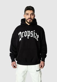 Svart hettegenser med en stor hvit "Dropsize"-logo, frontlomme og snor i hetten. Brukt med hvite cargo-bukser som har en liten logo.