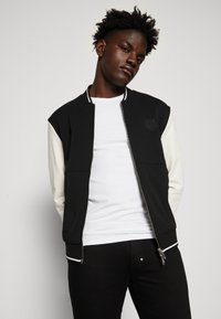 Chaqueta bomber negra y crema con cremallera frontal, cuello acanalado y un pequeño logo en el pecho. Usada sobre una camiseta blanca.