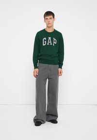 Grüner Pullover mit grauem 'GAP'-Logo, kombiniert mit lockeren grauen Jeans und schwarzen Schuhen. Einfaches Design, weicher Stoff, lässige Passform.