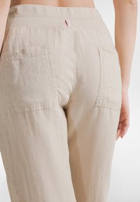 Beiget pellavahousut, joissa on leveä vyötärönauha, kaksi takataskua ja pieni merkkilappu. Kangas näyttää pehmeältä ja hienovaraisesti teksturoitulta.