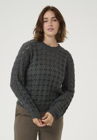 Maglione grigio in knit con una trama aperta e testurizzata. Collo rotondo, maniche lunghe e polsini a coste. Abbinato a pantaloni beige.