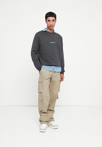 Sweatshirt gris foncé avec texte brodé, superposé à une chemise en denim bleu clair. Pantalons cargo crème avec plusieurs poches et sneakers blanches.