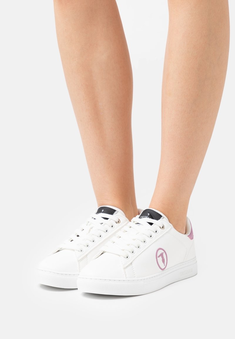 White sneakers with a smooth leather texture, round toe, black and pink accents on the heel and tongue, featuring a circular logo on the side.